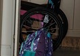 Photo is of an open door, propped by a backpack, with a wheelchair the other side.