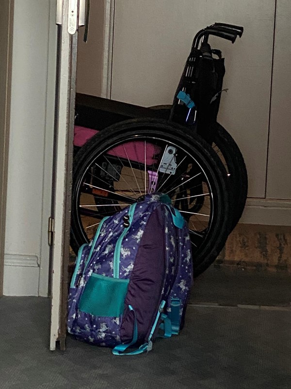 Photo is of an open door, propped by a backpack, with a wheelchair the other side.