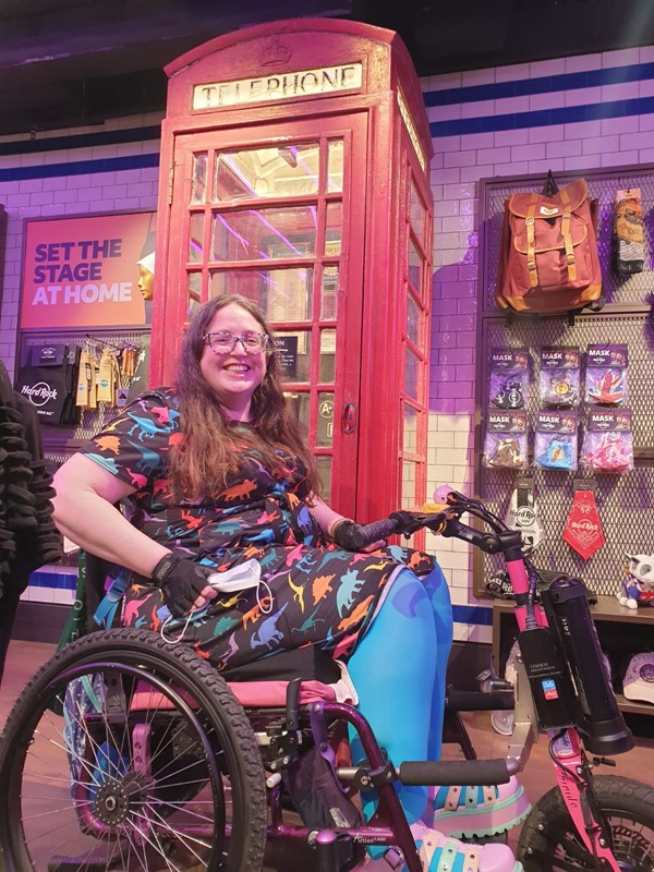 A white woman with long brown hair and glasses, sits in her wheelchair with power attachment. She wears a dress with dinosaurs on. She is sitting in front of a red telephone box.