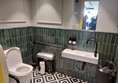 a view of the accessible toilet, showing the toilet, at a good height, with a rail on one side and a grab handle on the opposite wall. The sanitary bill and spare toilet rolls are within reach. The sink has space below to get close in a chair. The tap is easy to hold and operate. The soap is in reach. The mirror is at standing height.