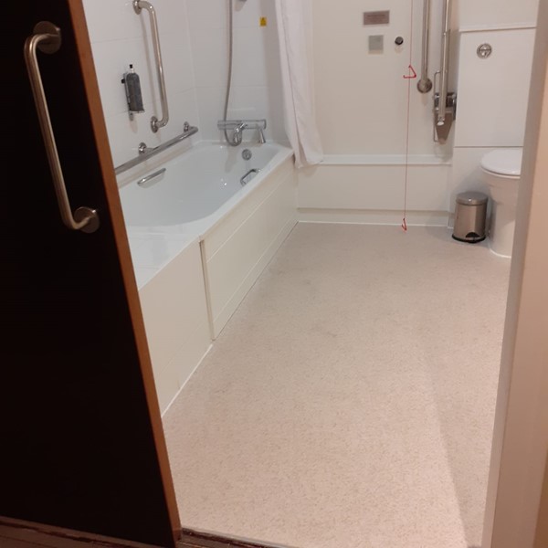 A view of the accessable bathroom from the door. There is a toilet with grab rails on both sides, a lowered bath tub with grab rails and a red emergency cord hanging to just above the floor.