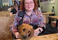 A white nonbinary woman with ginger curly hair, cat-eye glasses, and a pink and purple floral dress sits at a booth with her 16 week old golden retriever puppy assistance dog in training. Behind her is a green wall with two handwritten menus and a large sign that says "WILLIAM". In the background are other people at the cafe and a server.