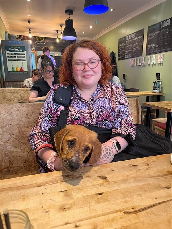 A white nonbinary woman with ginger curly hair, cat-eye glasses, and a pink and purple floral dress sits at a booth with her 16 week old golden retriever puppy assistance dog in training. Behind her is a green wall with two handwritten menus and a large sign that says "WILLIAM". In the background are other people at the cafe and a server.