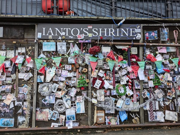 Image of the shrine, white letters on a black sign read "Ianto's Shrine" and there are plenty of pictures and tributes.
