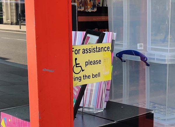 The entrance to the shop with a sign showing a wheelchair symbol and the words 'For assistance please ring the bell'. The doorbell button unit is missing.
