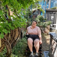 The image shows Pat, a white person with short brown hair wearing a pink shirt and black shorts. They are sitting in a powerchair and smiling at the camera. They are in a brick-paved yard surrounded by greenery.