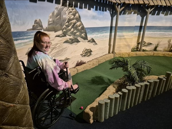 Nell in her wheelchair holding a golf club and looking back at the camera. In front of her is one of the holes at the golf course, displaying how bumpy it is in areas.