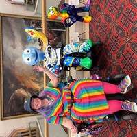 A photo of Vikki in their powerchair wearing a rainbow dress. They raise one arm with the other on their joystick to mimic the morph sculpture in a powerchair beside them.