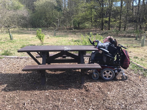Image of Claire sitting under the picnic bench to show that a wheelchair can fit underneath.