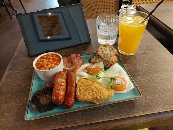The all day breakfast on the cafe table: sausages, bacon, hash browns, two free range eggs, a potrabello mushroom, beans and sourdough toast. Fresh squeezed orange juice and a glass of water. Claire's kindle is propped up behind the plate.