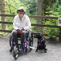 Lady in wheelchair with assistance dog sitting by her side. They are on a path with forest behind.