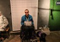 Lady in wheelchair with assistance dog, sitting in front of height chart. The lady and the dog are both holding prison number cards.