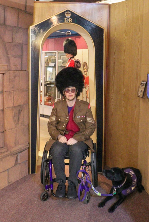 Lady in wheelchair wearing guard's jacket and hat.