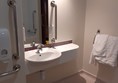 An accessable toilet showing a sink with clearance underneath for a wheelchair. There it's a mirror above the sink with a grab bar next to it. A towel rack is on the right wall.