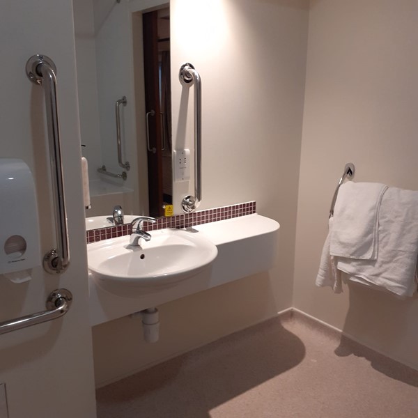 An accessable toilet showing a sink with clearance underneath for a wheelchair. There it's a mirror above the sink with a grab bar next to it. A towel rack is on the right wall.